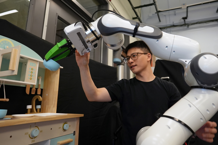 Graduate student Felix Yanwei Wang nudges a robotic arm that is manipulating a bowl in a toy kitchen set up in the group’s lab. Using the framework Wang and his collaborators developed, slightly nudging a robot is one way to correct its behavior (Credits: Melanie Gonick, MIT).